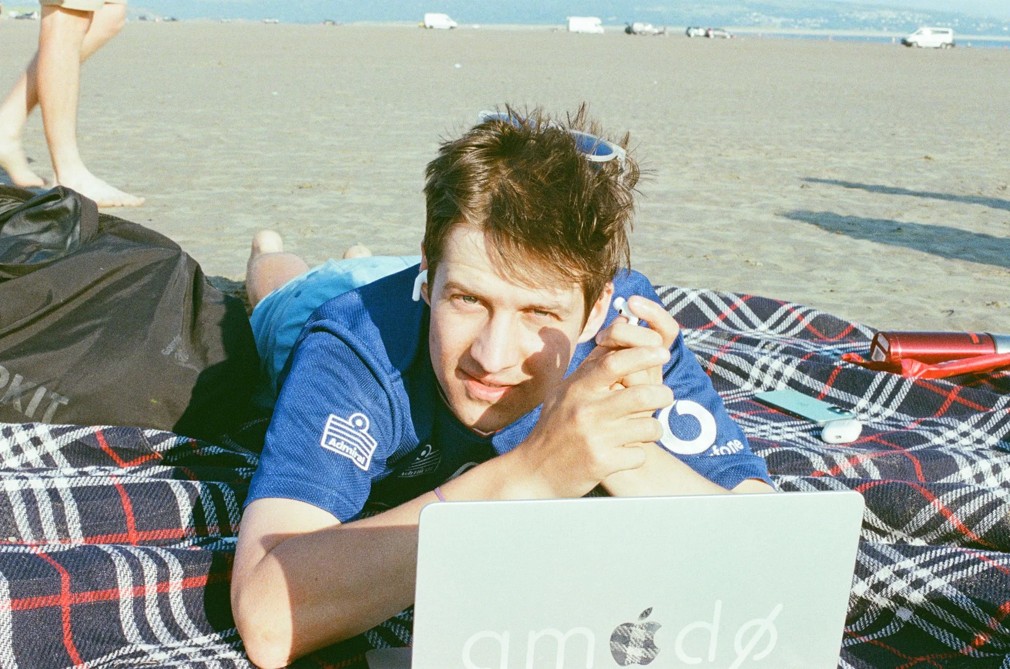 A man lying on a picnic blanket at the beach with a laptop